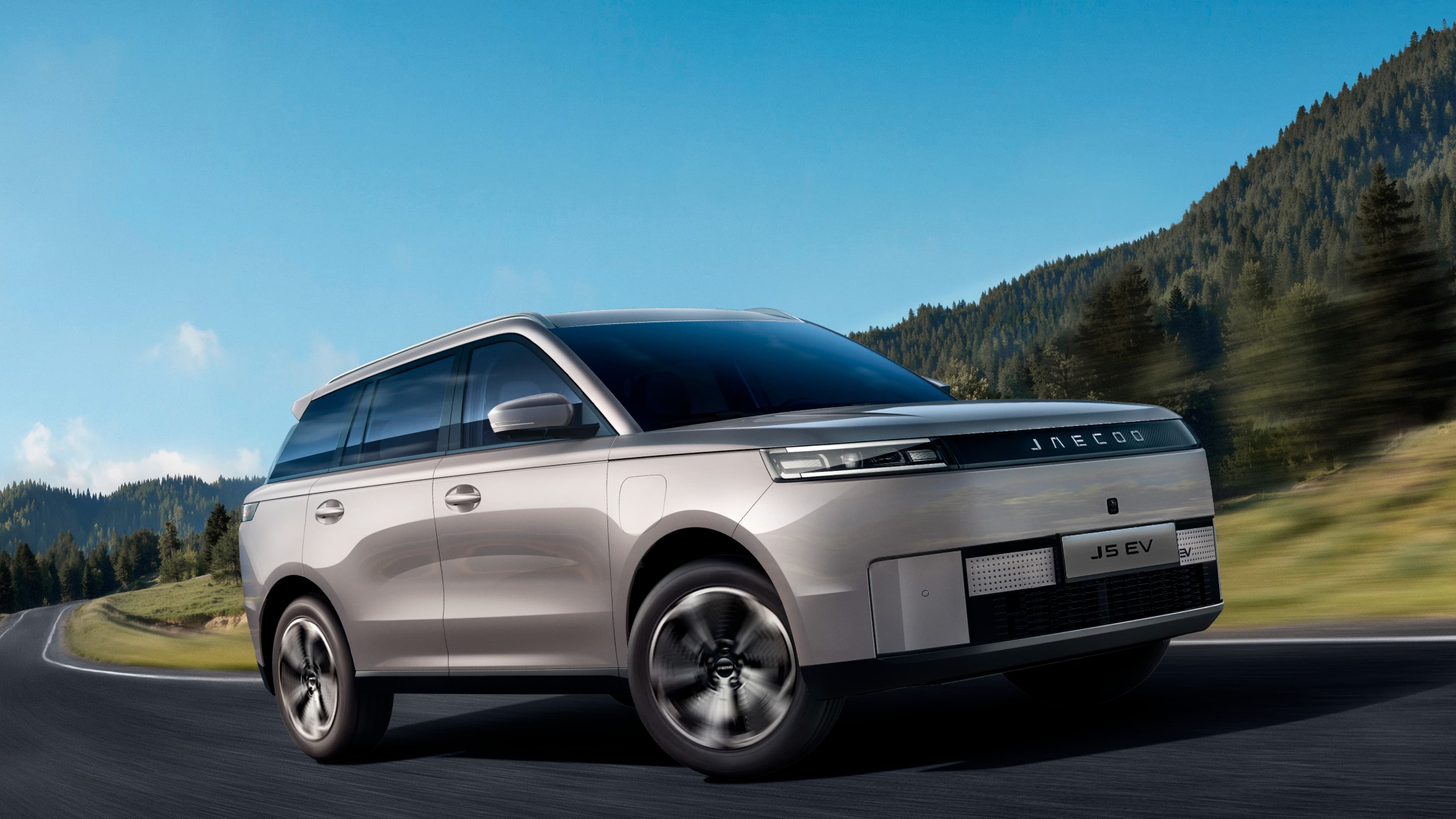 JAECOO J5 EV i sølv kører på en landevej med bjerge og græs i baggrunden på en dag med solskin.